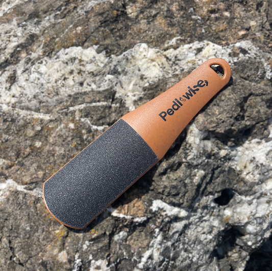 A double sided foot file with a brown handle and gray abrasive surface, placed on a rocky surface.